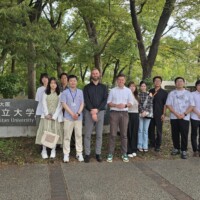 13 naukowców na tle drzew z prawej tabliczka z napisem Osaka Metropolitan University